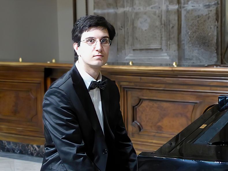 Na fotografii Aldo Roberto Pessolano, ubrany w czarny smoking, siedzi przy fortepianie.